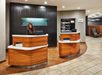 Hotel lobby with a smiling staff member behind a curved wooden reception desk, surrounded by modern decor with a snack shelf in the background.