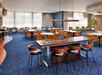 Spacious hotel lounge with wooden tables and leather chairs on blue carpet, featuring a large window and modern lighting.