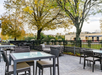 Outdoor patio with glass-top tables, wicker chairs, and trees with autumn leaves. Black metal fence surrounds the area, with a view of buildings and greenery in the background.