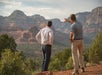 Two men stand on a rocky overlook surrounded by trees, facing and looking out at red rock formations and distant mountains. One man gestures towards the landscape.