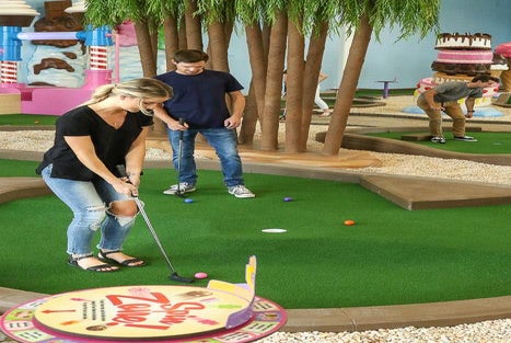 Three people play mini-golf on an indoor course with artificial grass, surrounded by playful decor like trees and oversized candy props.