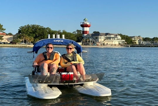 Two people wearing life jackets sit on a small pontoon boat on calm water, with a lighthouse and buildings visible in the background.