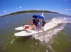 Two people ride a small motorized catamaran on a sunny day, wearing life jackets, with water splashing around the boat and land visible in the background.