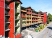 A multi-story red and green apartment building with exterior staircases, adjacent to a tree-lined street.