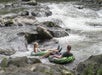 Two people float on inflatable tubes down a rocky river, navigating through gentle rapids surrounded by rocks and flowing water.