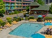 Outdoor pool area with people lounging on yellow chairs. A colorful multi-story building and a green-roofed pavilion are in the background.