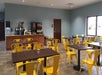 A small dining area with several dark wooden tables, each surrounded by bright yellow metal chairs. A beverage station is located in the background against a wall.