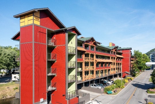 A colorful, multi-story building with balconies and a peaked roof, set beside a road and trees under a clear blue sky.