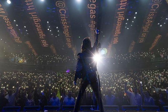 A performer stands on stage facing a large audience holding up phone lights, with bright stage lighting and social media handles projected on the ceiling.