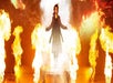 A person stands on a stage surrounded by fire and smoke, arms outstretched, wearing a long coat.