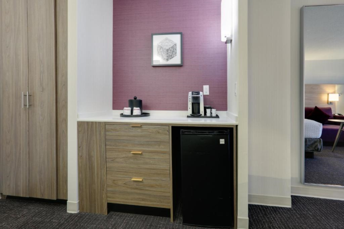 A hotel room kitchenette with wood cabinets, a black mini fridge, a coffee maker, ice bucket, and cups on a counter, with a purple accent wall and framed artwork above.
