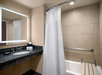 Hotel bathroom with a granite countertop, sink, and mirror. Includes a bathtub with a white shower curtain and a wooden bench. A grab bar is mounted on the wall.
