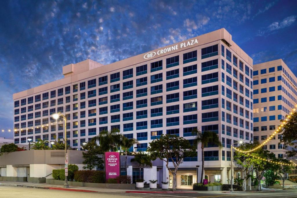 Exterior view of Crowne Plaza hotel at dusk, with illuminated sign on top, surrounded by palm trees and street lights against a cloudy sky.