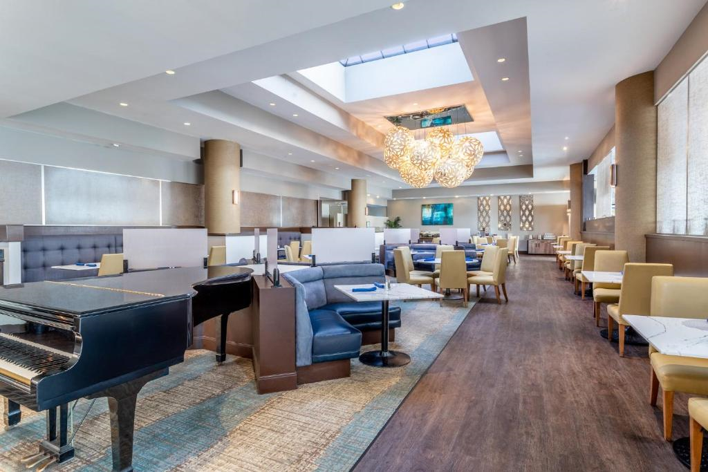 Spacious restaurant interior with modern decor, featuring a central chandelier, multiple dining tables, and a grand piano on a wooden floor. Ceiling features skylights.