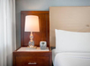 A bedside table with a lamp, digital clock, and drawer next to a bed with white bedding.