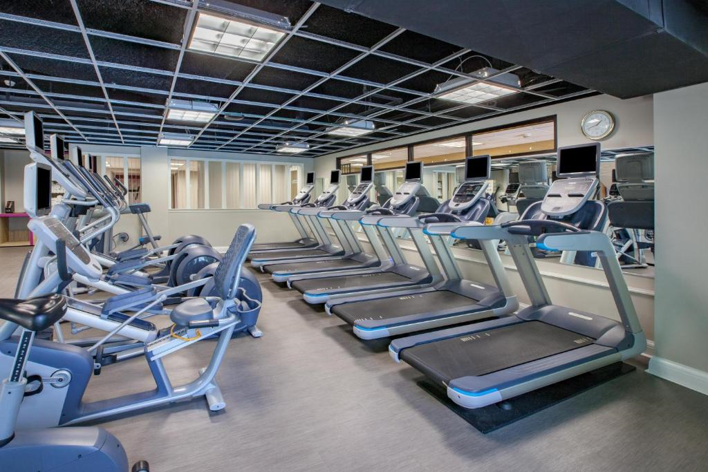 A modern gym features a row of treadmills and stationary bikes on a gray floor, with mirrors and a clock on the wall. The ceiling has a grid pattern with fluorescent lights.