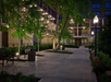 Paved walkway with benches and street lamps runs through a landscaped courtyard surrounded by multi-story buildings at dusk.