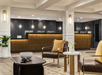 Modern hotel lobby with seating area in the foreground and a front desk with four computer stations in the background. Neutral colors and contemporary decor are featured throughout.