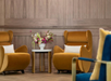 Two mustard yellow armchairs with white pillows flank a small round table holding a vase of flowers, set against a wood-paneled wall.