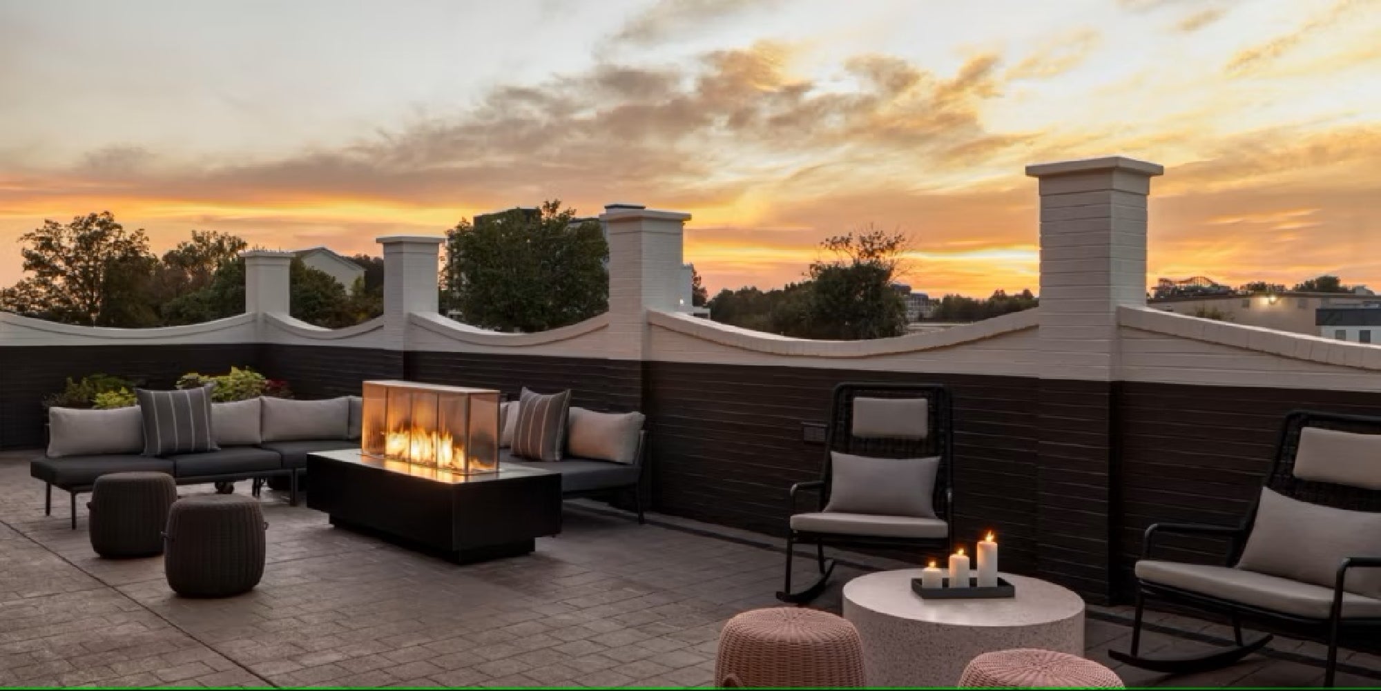 A rooftop patio at sunset features cushioned seating, a modern fire pit, and a small table with candles, enclosed by a decorative fence.