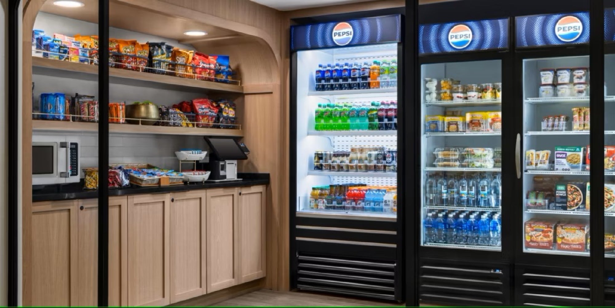 A self-serve convenience store area with shelves of snacks, a microwave, drink refrigerators, and a checkout counter with a register.