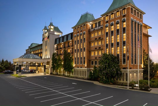 Large brick hotel with multiple stories and green roofs, illuminated at dusk, with an empty parking lot and main entrance visible.