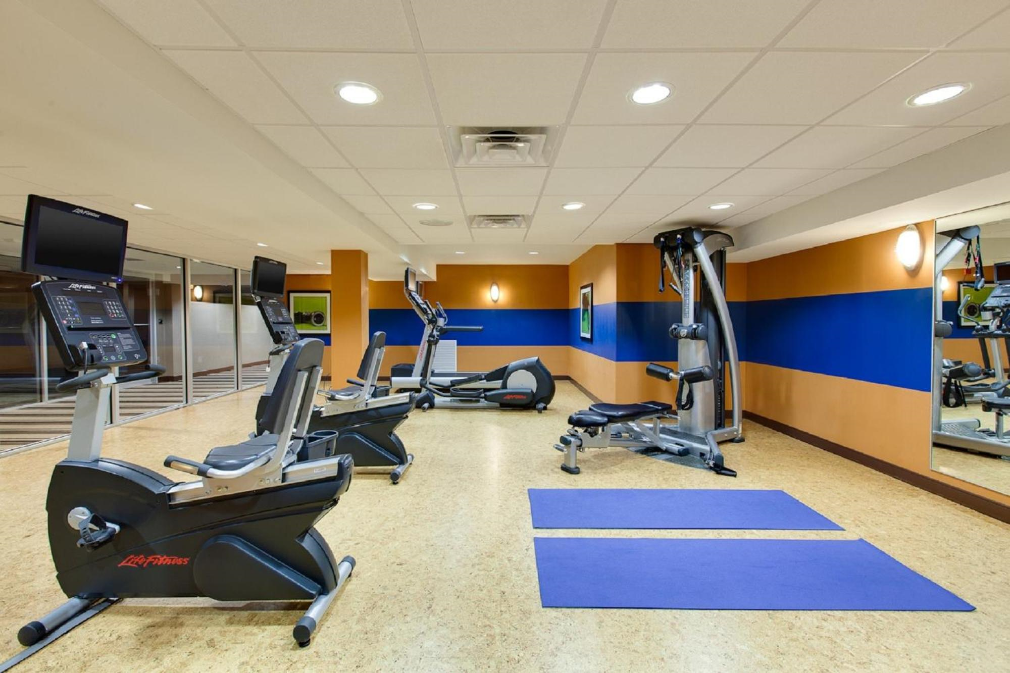 A gym room with exercise bikes, a weight machine, two blue yoga mats, wall mirrors, and brightly colored walls under ceiling lights.