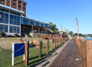 Wooden boardwalk and grassy area beside a waterfront restaurant with outdoor seating on a sunny day.