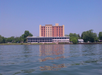 A multi-story hotel building with a flat roof is seen from across a calm body of water, surrounded by trees under a clear blue sky.