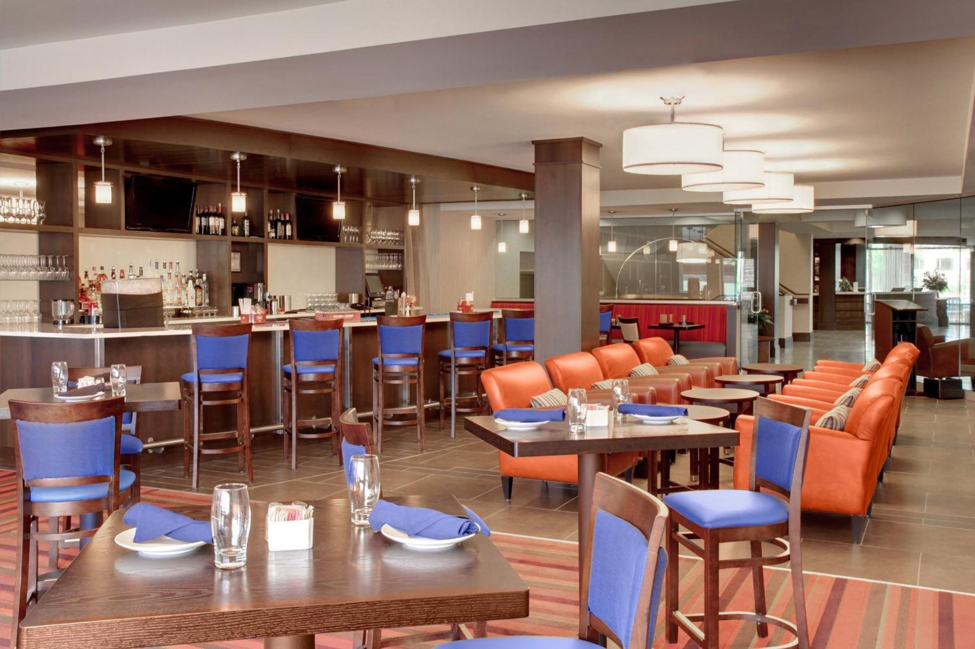 Modern restaurant interior with wooden tables, blue chairs, orange armchairs, a bar area with stools, and hanging white lights. Tables are set with glassware and napkins.