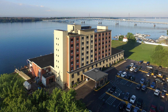 A multi-story hotel building stands near a river with a long bridge in the background, adjacent to a parking lot and a marina, surrounded by greenery.