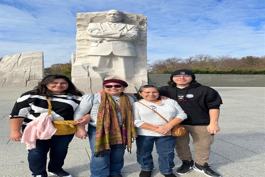 Four people stand in front of a tall stone monument depicting a figure with crossed arms on a clear day.