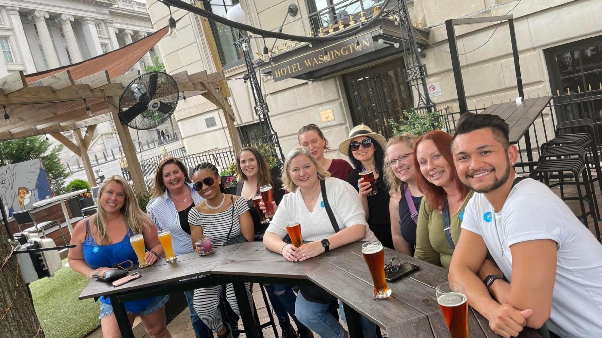 A group of people sit and smile at an outdoor table with drinks in front of Hotel Washington on a cloudy day.