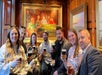 Seven people sit indoors holding pints of beer, smiling at the camera, with a large painting of card players hanging on the wood-paneled wall behind them.