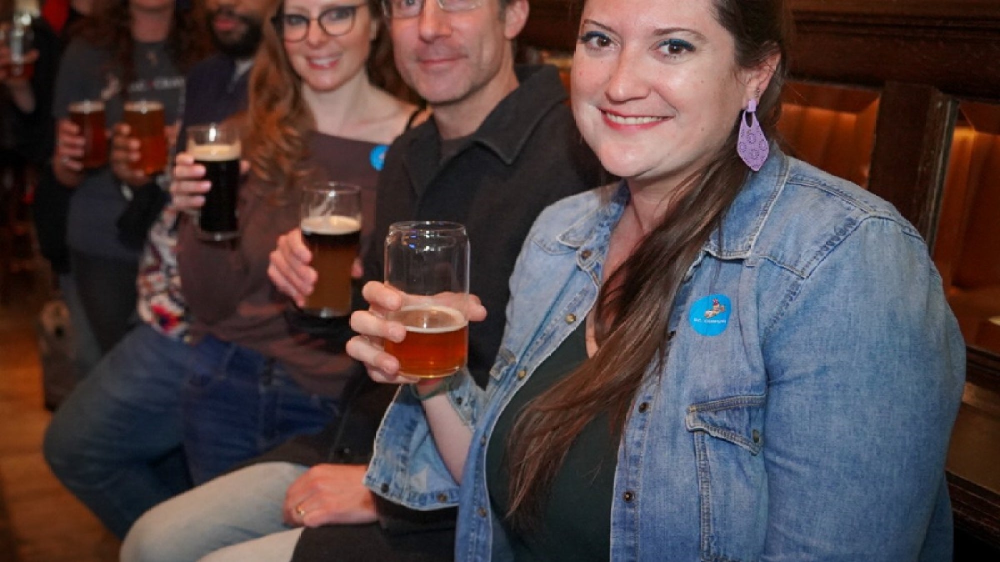 A group of people sit side by side in a bar, holding glasses of beer and smiling at the camera.