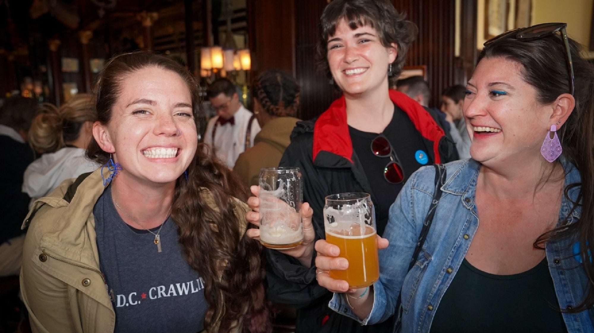 Three women are standing close together indoors, smiling and holding glasses of beer in a lively, crowded bar or pub setting.