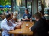 A man shows a tablet to five people seated around a wooden table with drinks, inside a restaurant with large windows overlooking an outdoor patio.