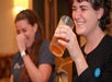 Two women sitting indoors, laughing, with one holding a glass of amber-colored drink and the other covering her mouth.