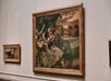 A framed painting of dancers in a scenic landscape hangs on a gallery wall next to another smaller painting.