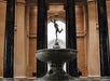 A large stone fountain with a bronze statue at the center is surrounded by tall marble columns in an ornate interior space.