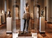 A bronze ballerina statue wearing a tutu is displayed in a museum gallery surrounded by other sculptures.