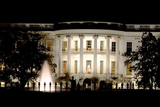 The White House illuminated at night, with lights on in some windows and a water fountain in the front yard.
