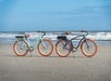 Two beach cruiser bicycles with tan tires, one teal and one navy, are parked side by side on a sandy beach with the ocean waves in the background.