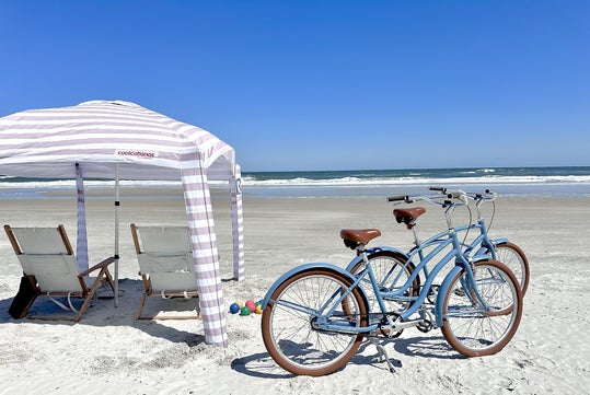 Two blue bicycles parked on a sandy beach beside a striped canopy with two chairs under it. Ocean waves are visible in the background under a clear blue sky.