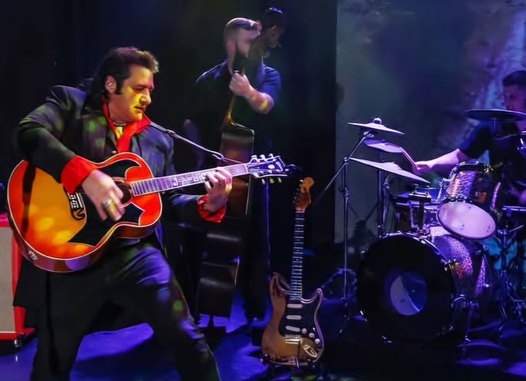 A guitarist performs energetically on stage with a double bassist and a drummer; an electric guitar is placed on a stand nearby.