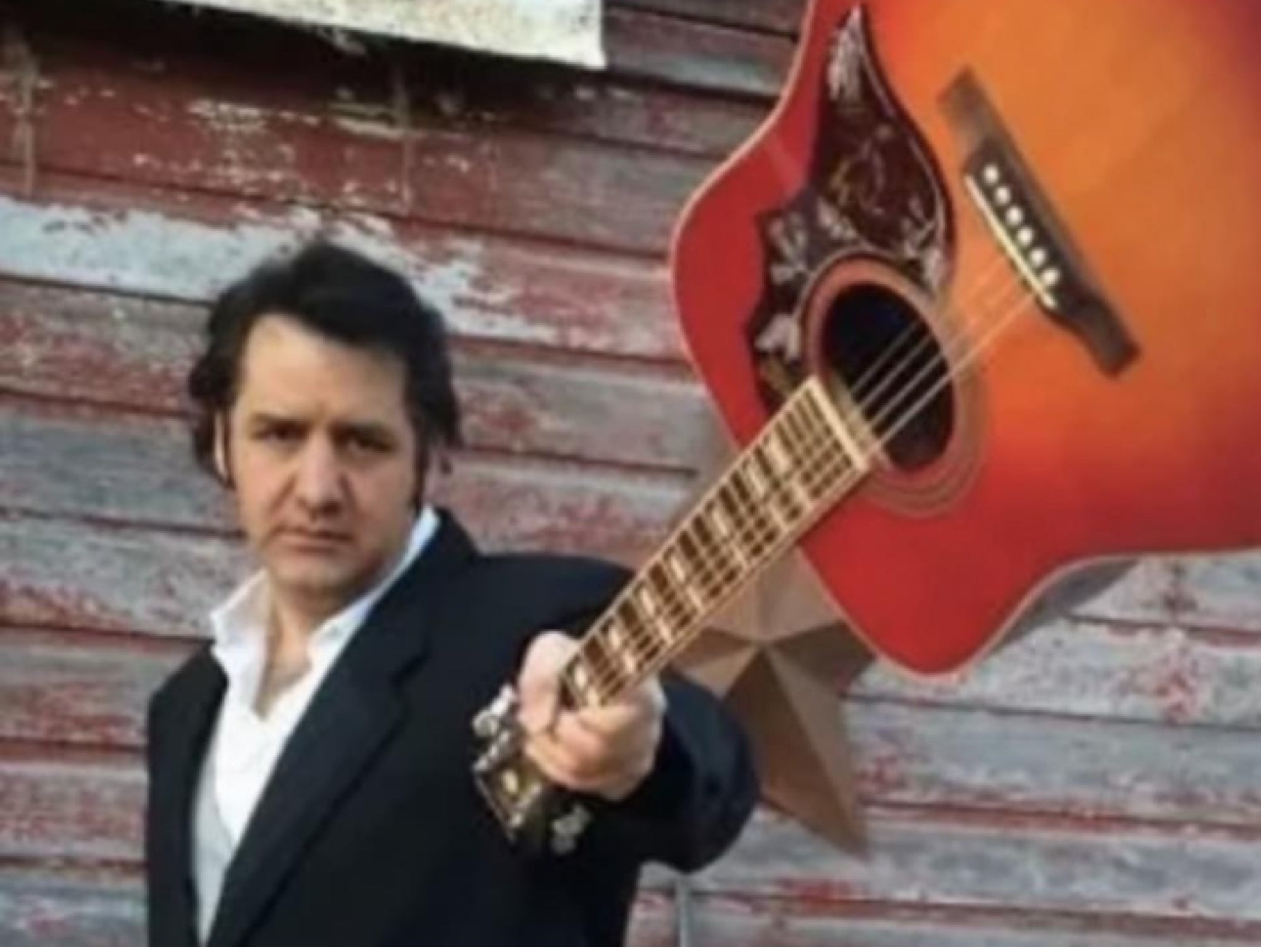 A man in a dark suit holds an acoustic guitar by the neck, extending it outward, standing in front of a weathered wooden wall.