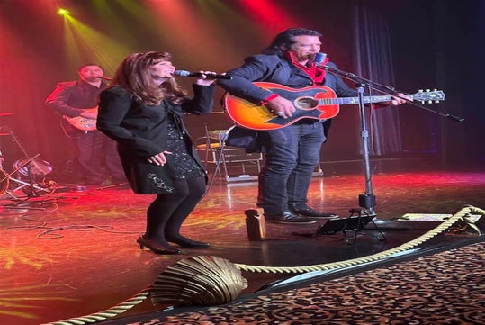 A man plays guitar and sings into a microphone beside a woman also singing on stage, with a band performing in the background under stage lights.
