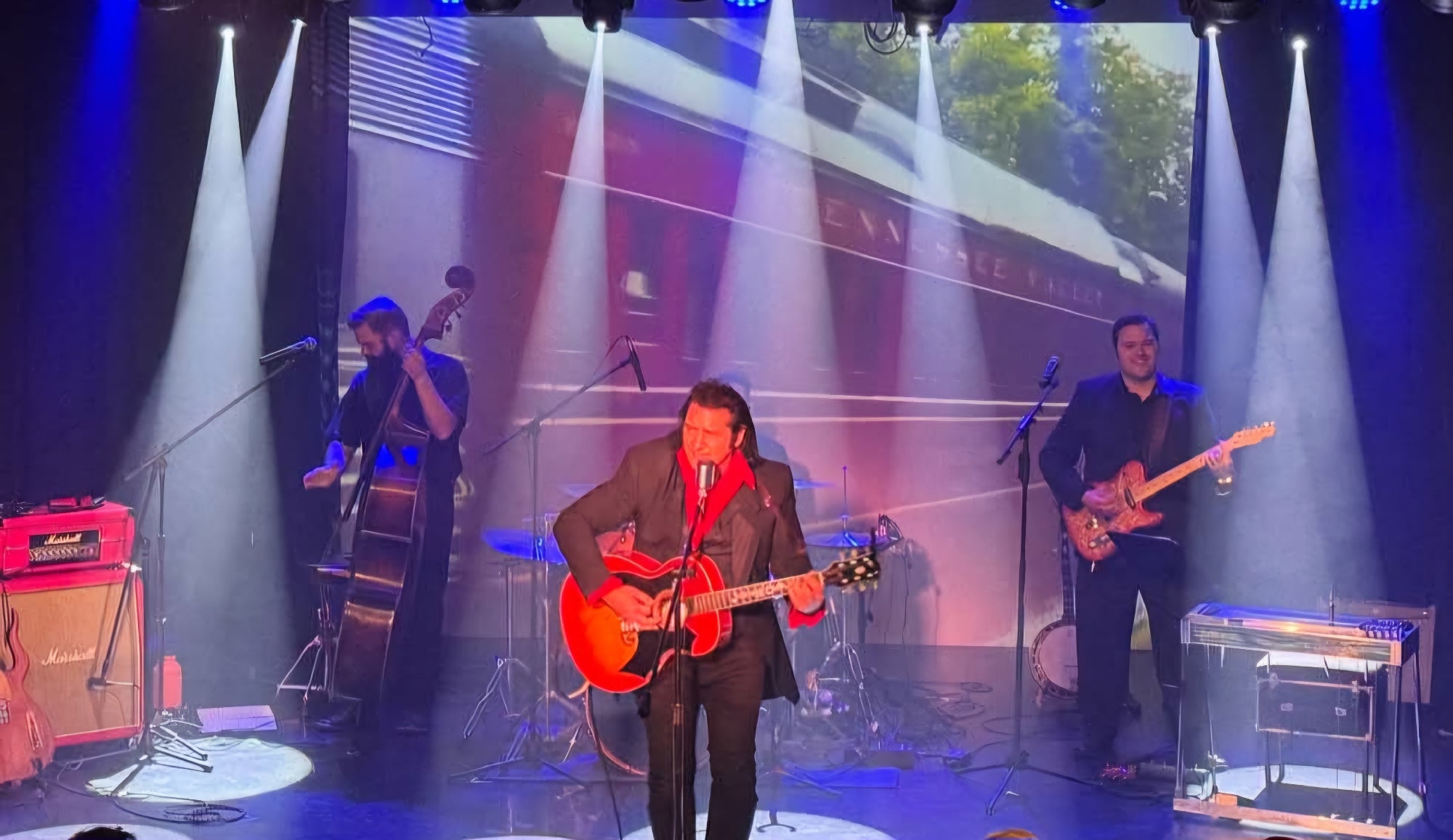 A four-piece band performs on stage with guitars and a double bass, while a video of a moving train is projected on a screen behind them under blue stage lights.