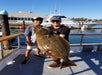 Two anglers proudly displaying a halibut catch on the deck of a boat at the dock, with another large fishing vessel in the background.