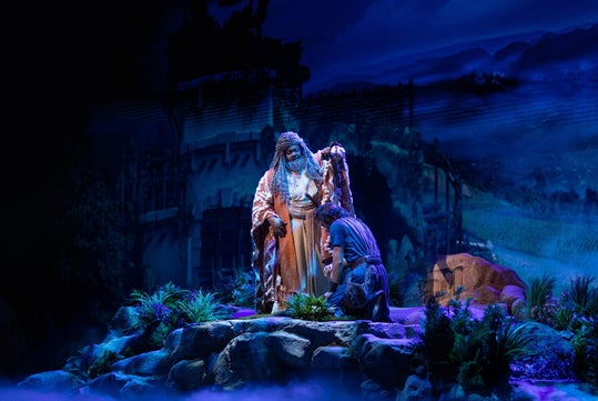 Stage scene with two people in dramatic lighting. One person stands in elaborate robes, the other kneels in front. The background depicts a dark, mountainous landscape with structures.